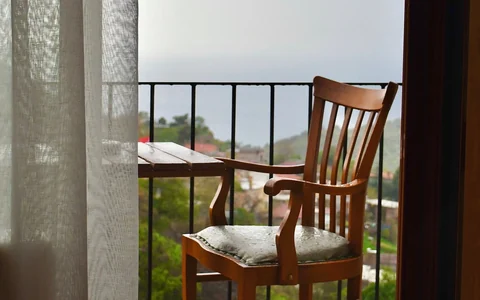 Cadeira e mesa em varanda vista através de porta e cortina; fotografia ilustrativa, sem vínculo com oferta local.